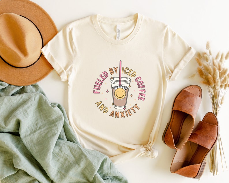 May include: A white t-shirt with a graphic design of a cup of iced coffee with a smiley face and the text "Fueled by Iced Coffee and Anxiety".