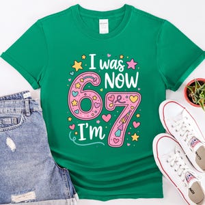 May include: A green t-shirt featuring the text "I was 67, Now I'm 67" in a colourful design. The design includes pink numbers, stars, hearts, and a party hat. The shirt is shown with denim shorts and white trainers.