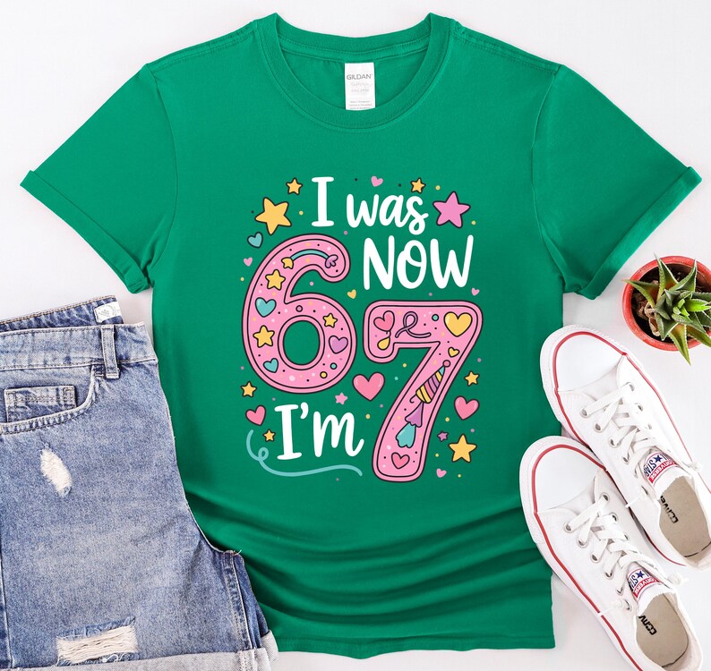 May include: A green t-shirt featuring the text "I was 67, Now I'm 67" in a colourful design. The design includes pink numbers, stars, hearts, and a party hat. The shirt is shown with denim shorts and white trainers.