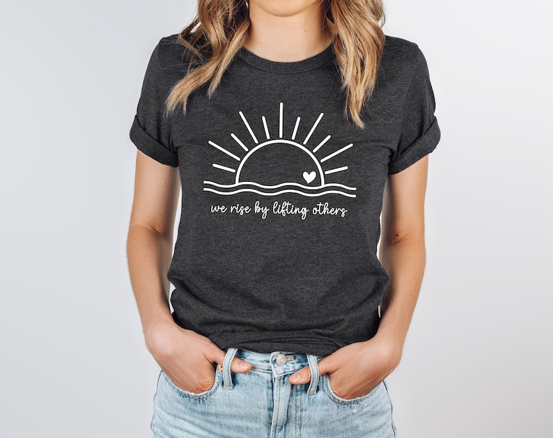 K&ouml;nnte beinhalten: Ein dunkelgraues T-Shirt mit einer wei&szlig;en Grafik einer Sonne, die &uuml;ber einer Welle mit einem Herzen in der Mitte aufgeht. Der Text "we rise by lifting others" ist unter der Grafik gedruckt.