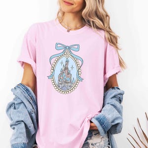 Puede incluir: Una camiseta rosa con una ilustraci&oacute;n azul y blanca de un castillo con un lazo azul en la parte superior. El castillo est&aacute; rodeado por un borde de perlas.