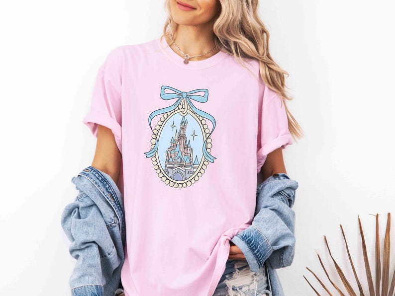 Puede incluir: Una camiseta rosa con una ilustraci&oacute;n azul y blanca de un castillo con un lazo azul en la parte superior. El castillo est&aacute; rodeado por un borde de perlas.