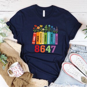 May include: Navy blue t-shirt featuring a colorful design. The design includes a row of books with words like "Love", "Peace", and "Justice", topped with flowers. Below the books is the number "8647" in rainbow colors. The shirt is paired with jeans and white sneakers.