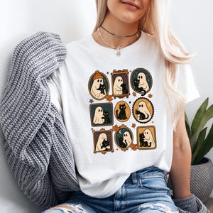Puede incluir: Camiseta blanca con un dise&ntilde;o tem&aacute;tico de Halloween. El gr&aacute;fico incluye ilustraciones enmarcadas de fantasmas y gatos negros, con hojas de oto&ntilde;o y una paleta de colores c&aacute;lidos. Una prenda casual para el d&iacute;a a d&iacute;a.