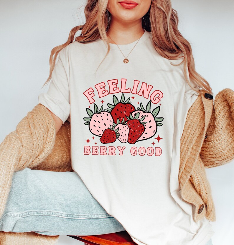 May include: A white t-shirt with a pink and red graphic of strawberries and the text "Feeling Berry Good".