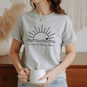 K&ouml;nnte beinhalten: Ein graues T-Shirt mit einer schwarzen Grafik einer Sonne, die &uuml;ber einer Welle mit einem Herzen in der Sonne aufgeht. Der Text "we rise by lifting others" befindet sich unter der Grafik.