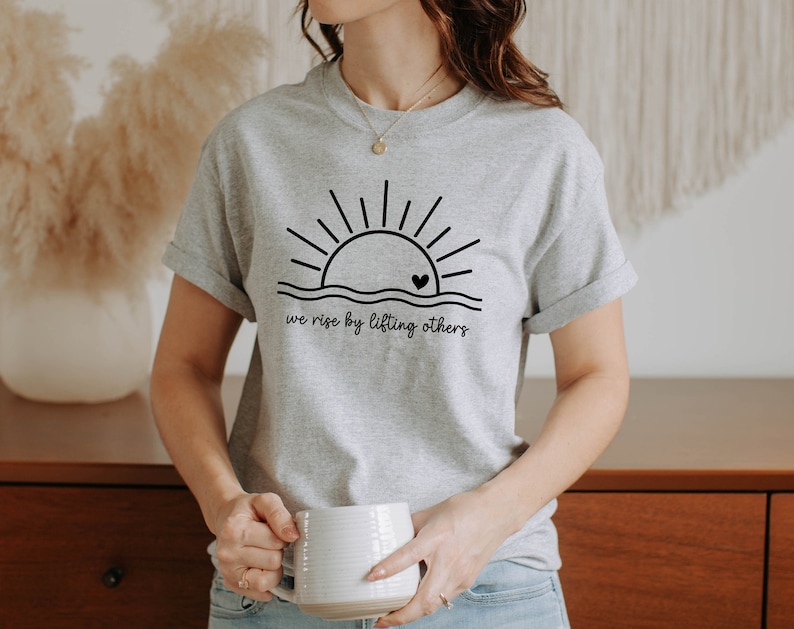 K&ouml;nnte beinhalten: Ein graues T-Shirt mit einer schwarzen Grafik einer Sonne, die &uuml;ber einer Welle mit einem Herzen in der Sonne aufgeht. Der Text "we rise by lifting others" befindet sich unter der Grafik.