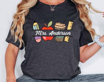 May include: Dark gray t-shirt featuring a colorful graphic design for a teacher. The design includes a pencil, apple, coffee cup, ruler, and the name "Mrs. Anderson" in white text. The shirt is made of a soft material.