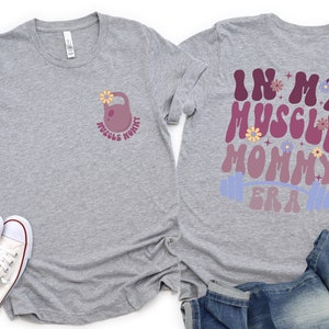 Op de afbeelding: Grijs T-shirt met een roze halter met een bloem erop en de tekst "Muscle Mommy" aan de voorkant. De achterkant van het shirt heeft de tekst "In My Muscle Mommy Era" in een retro lettertype met een roze en paarse gradi&euml;nt en een blauwe halter.