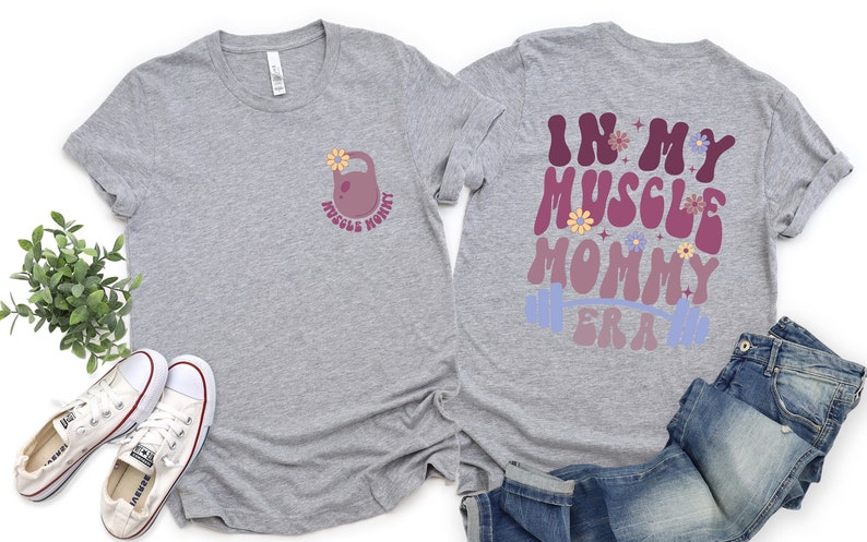 Op de afbeelding: Grijs T-shirt met een roze halter met een bloem erop en de tekst "Muscle Mommy" aan de voorkant. De achterkant van het shirt heeft de tekst "In My Muscle Mommy Era" in een retro lettertype met een roze en paarse gradi&euml;nt en een blauwe halter.