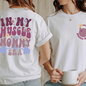 Op de afbeelding: Wit T-shirt met de tekst "In My Muscle Mommy Era" in een retro lettertype met bloemen en een halterstang op de rug. De voorkant van het shirt heeft een paarse kettlebell met een bloem erop en de tekst "Muscle Mommy" eromheen.