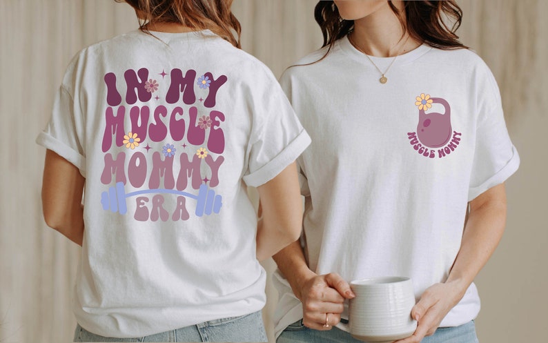 Op de afbeelding: Wit T-shirt met de tekst "In My Muscle Mommy Era" in een retro lettertype met bloemen en een halterstang op de rug. De voorkant van het shirt heeft een paarse kettlebell met een bloem erop en de tekst "Muscle Mommy" eromheen.