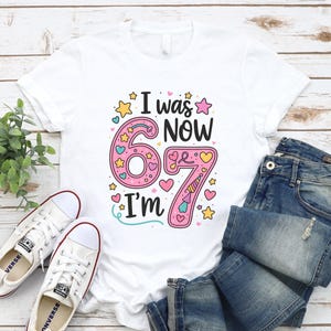 May include: White t-shirt with the text "I was 67, now I'm 67" in a colourful design. The number 67 is pink with hearts and stars. The shirt is paired with blue jeans and white trainers.
