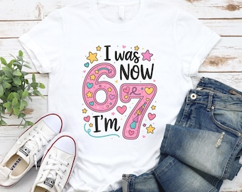May include: White t-shirt with the text "I was 67, now I'm 67" in a colorful design. The number 67 is pink with hearts and stars. The shirt is paired with blue jeans and white sneakers.