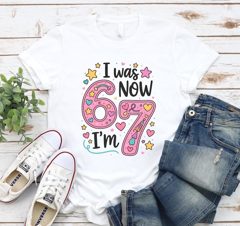 May include: White t-shirt with the text "I was 67, now I'm 67" in a colourful design. The number 67 is pink with hearts and stars. The shirt is paired with blue jeans and white trainers.