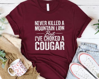 May include: A maroon t-shirt with the text "NEVER KILLED A MOUNTAIN LION But I'VE CHOKED A COUGAR" in white, distressed lettering. The shirt is folded, with a coffee mug and sneakers visible.