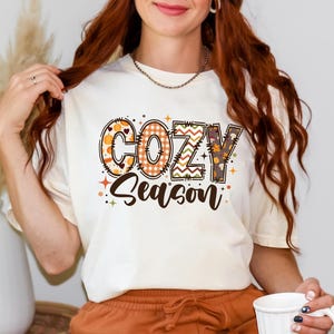 May include: Cream-colored t-shirt with the words "COZY Season" in a colorful, fall-themed design. The letters are decorated with patterns like pumpkins, plaid, and leaves. The shirt is a casual, seasonal apparel item.