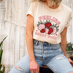 May include: A cream colored t-shirt with a graphic of strawberries and the text "Feeling Berry Good" in pink and red lettering.