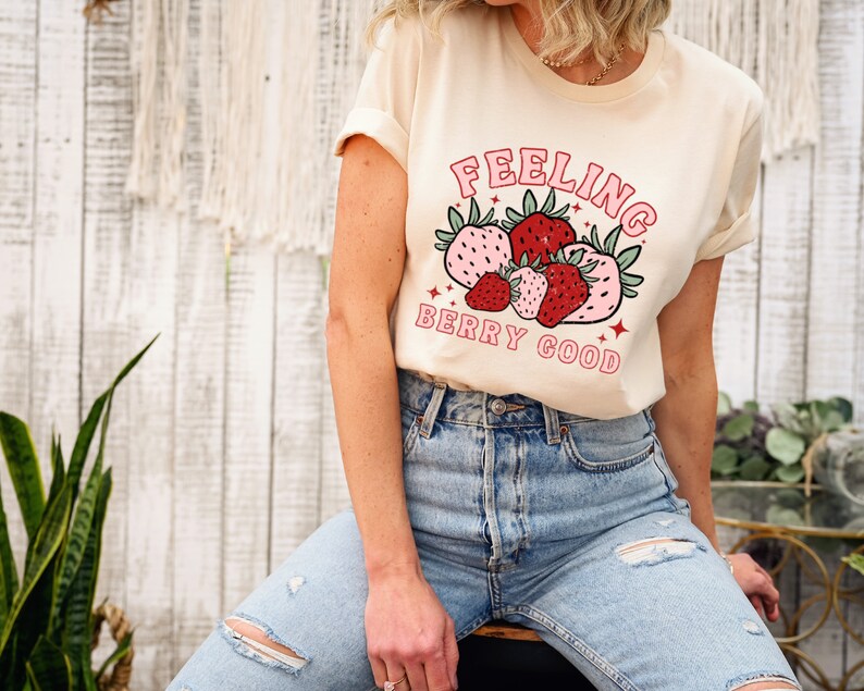 May include: A cream colored t-shirt with a graphic of strawberries and the text "Feeling Berry Good" in pink and red lettering.