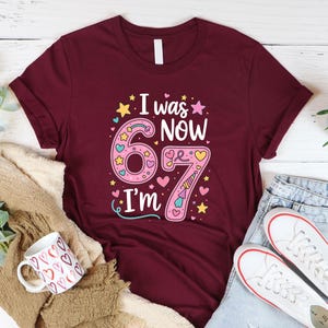 May include: Burgundy t-shirt with the text "I was NOW 67 I'm" in a colourful design. The number 6 is pink with hearts and stars, and the number 7 is pink with a unicorn horn. A white coffee mug with heart designs and white trainers are also visible.