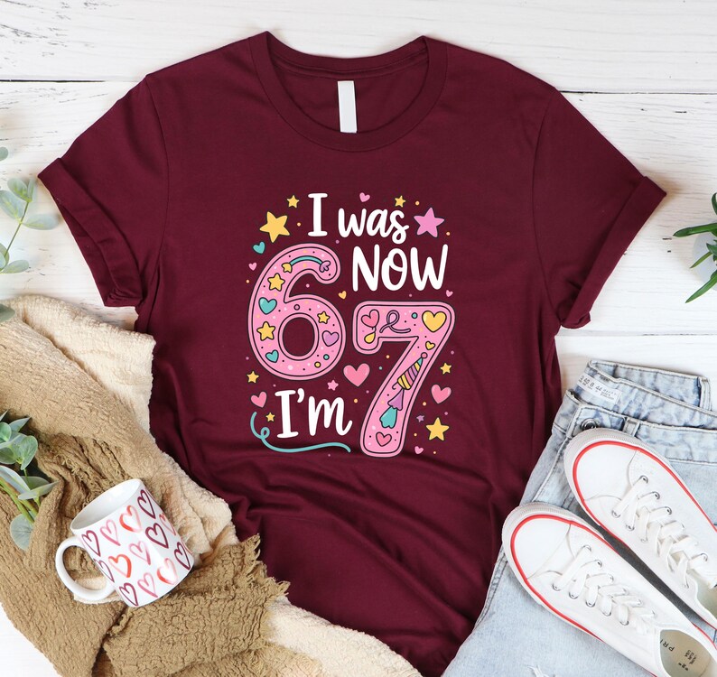 May include: Burgundy t-shirt with the text "I was NOW 67 I'm" in a colourful design. The number 6 is pink with hearts and stars, and the number 7 is pink with a unicorn horn. A white coffee mug with heart designs and white trainers are also visible.