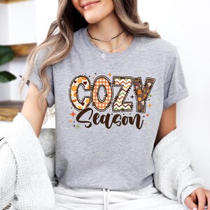 May include: A gray t-shirt with the words "COZY Season" in a decorative, fall-themed design. The letters feature patterns like pumpkins, plaid, and leaves. The shirt is a casual, everyday wear item.