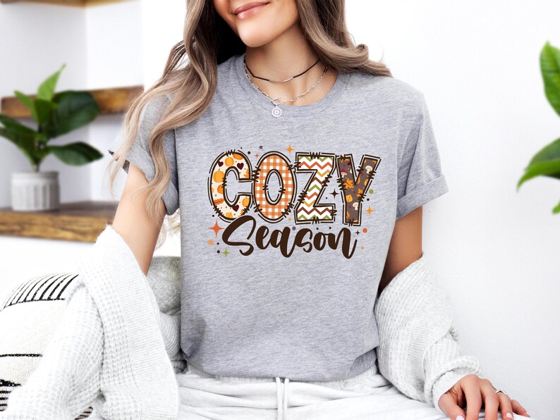 May include: A gray t-shirt with the words "COZY Season" in a decorative, fall-themed design. The letters feature patterns like pumpkins, plaid, and leaves. The shirt is a casual, everyday wear item.