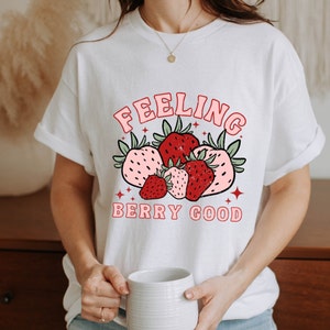 May include: White t-shirt with a red and pink strawberry graphic and the text "Feeling Berry Good".