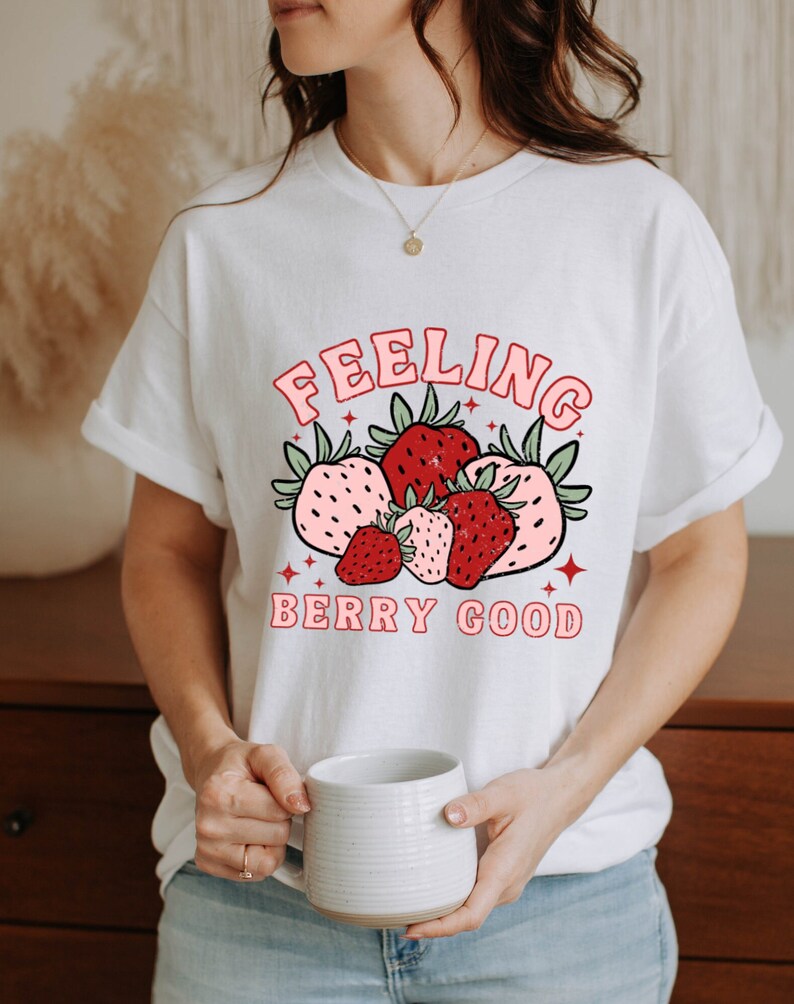 May include: White t-shirt with a red and pink strawberry graphic and the text "Feeling Berry Good".