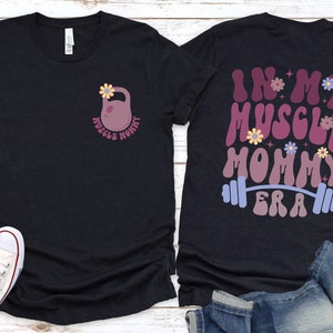 Op de afbeelding: Twee zwarte T-shirts met een fitness thema. Het voorste shirt heeft een roze kettlebell met een bloem erop en de tekst "Muscle Mommy". Het achterste shirt heeft de tekst "In My Muscle Mommy Era" in een retro lettertype met een halterstang en bloemen.