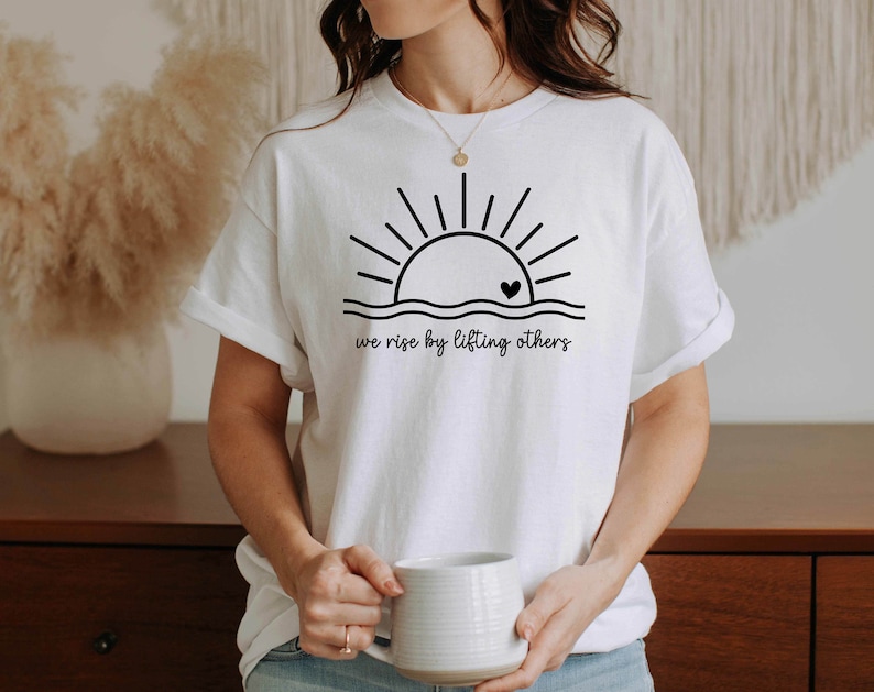 K&ouml;nnte beinhalten: Wei&szlig;es T-Shirt mit einer schwarzen Grafik einer Sonne, die &uuml;ber Wellen mit einem Herzen in der Mitte aufgeht. Der Text "we rise by lifting others" befindet sich unter der Grafik.