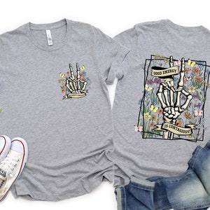 May include: Grey t-shirt with a floral design and a skeleton hand making a peace sign. The text on the shirt reads "Good energy is contagious".