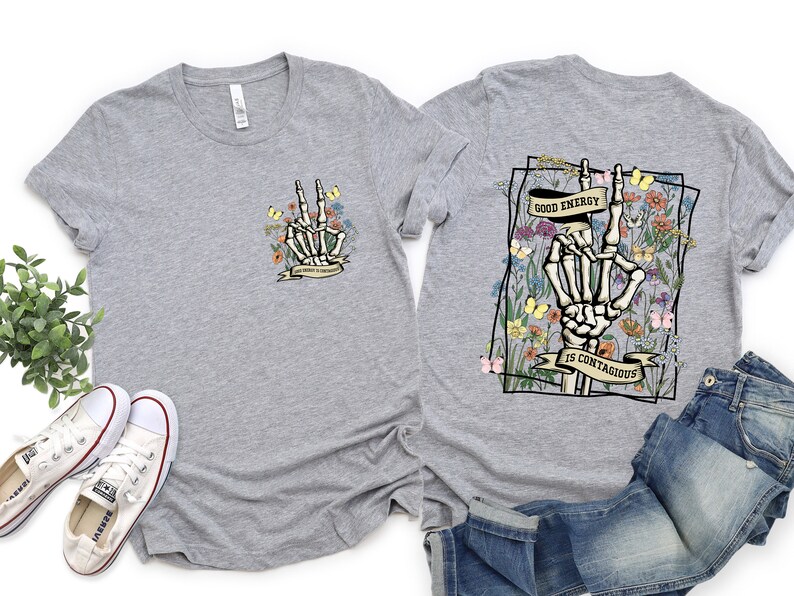 May include: Grey t-shirt with a floral design and a skeleton hand making a peace sign. The text on the shirt reads "Good energy is contagious".