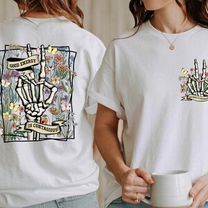 May include: White t-shirt featuring a graphic print of a skeleton hand making a peace sign surrounded by flowers and butterflies. The text "Good Energy is Contagious" is printed on a banner below the hand.