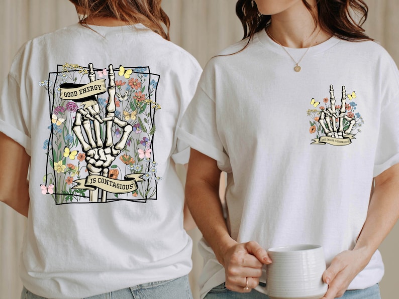 May include: White t-shirt featuring a graphic print of a skeleton hand making a peace sign surrounded by flowers and butterflies. The text "Good Energy is Contagious" is printed on a banner below the hand.