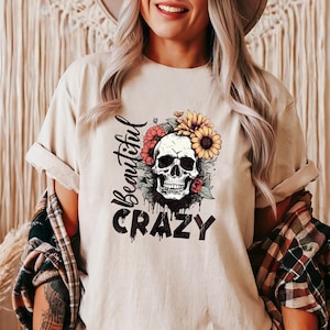 May include: Beige t-shirt with a skull graphic surrounded by flowers. The words "Beautiful Crazy" are printed in black script and block letters. The shirt has rolled-up sleeves.