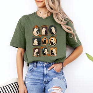 Puede incluir: Camiseta verde oliva con un gr&aacute;fico enmarcado de fantasmas y gatos negros. El dise&ntilde;o incluye fantasmas con sombreros y sosteniendo gatos, con hojas de oto&ntilde;o y detalles de luna. La camiseta es de estilo casual.