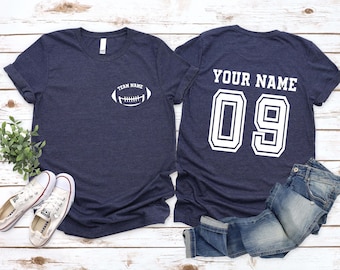 Könnte beinhalten: Zwei marineblaue T-Shirts mit weißen Grafiken. Eines hat eine Football-Grafik und den Text "TEAM NAME". Das andere hat "YOUR NAME" und die Nummer "09". Weiße Turnschuhe und eine blaue Jeans sind ebenfalls im Bild zu sehen.