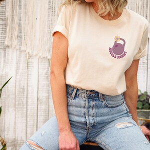 Op de afbeelding: Een licht beige t-shirt met een afbeelding van een paarse kettlebell met een gele bloem erop. De tekst "Muscle Mommy" staat rond de kettlebell.