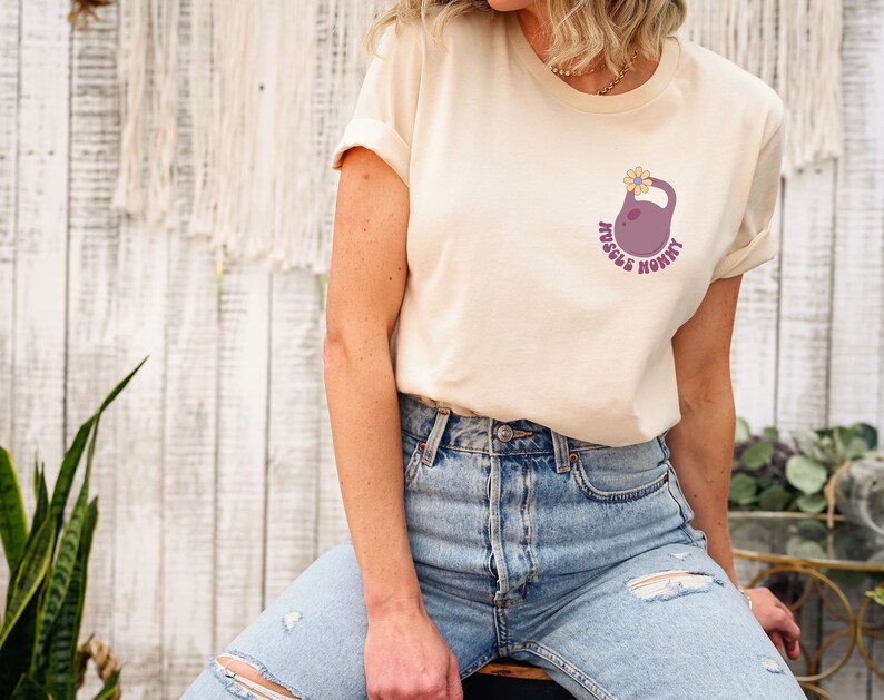 Op de afbeelding: Een licht beige t-shirt met een afbeelding van een paarse kettlebell met een gele bloem erop. De tekst "Muscle Mommy" staat rond de kettlebell.