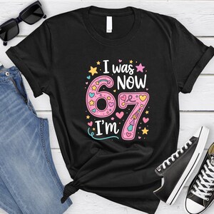 May include: Black t-shirt with the text "I was 67, now I'm 67" in pink, yellow, and blue. The number 6 is decorated with stars and hearts, and the number 7 has a unicorn horn. The shirt is paired with jeans and black trainers.