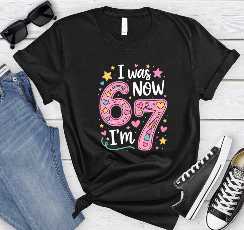 May include: Black t-shirt with the text "I was 67, now I'm 67" in pink, yellow, and blue. The number 6 is decorated with stars and hearts, and the number 7 has a unicorn horn. The shirt is paired with jeans and black trainers.