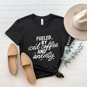 May include: Black t-shirt with white text that reads "Fueled by iced coffee and anxiety". The shirt is laid flat on a white surface with a pair of brown flats and a brown hat.