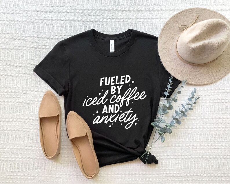 May include: Black t-shirt with white text that reads "Fueled by iced coffee and anxiety". The shirt is laid flat on a white surface with a pair of brown flats and a brown hat.