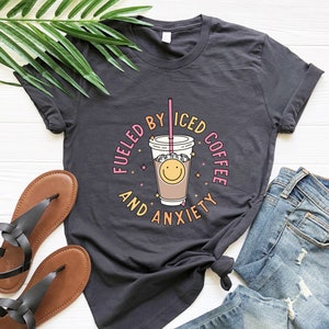 May include: A dark gray t-shirt with a graphic of a smiling iced coffee cup and the text "Fueled by Iced Coffee and Anxiety".
