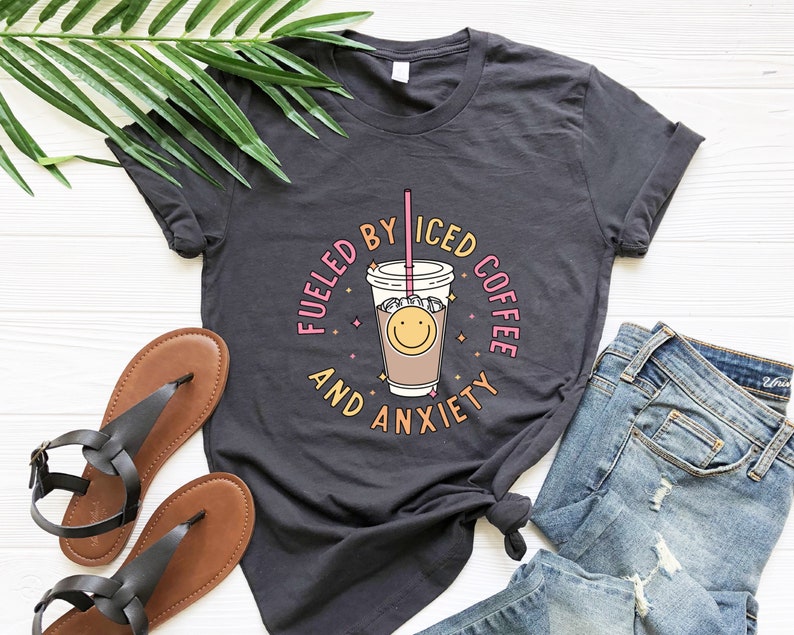 May include: A dark gray t-shirt with a graphic of a smiling iced coffee cup and the text "Fueled by Iced Coffee and Anxiety".