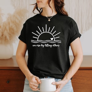 K&ouml;nnte beinhalten: Schwarzes T-Shirt mit einem wei&szlig;en Grafikdruck eines Sonnenaufgangs mit einem Herzen in der Mitte und dem Text "we rise by lifting others".