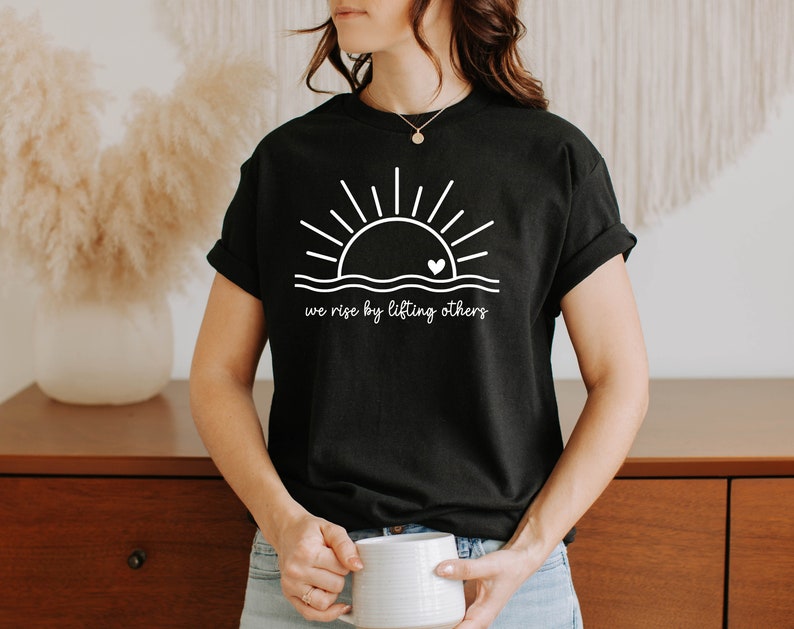 K&ouml;nnte beinhalten: Schwarzes T-Shirt mit einem wei&szlig;en Grafikdruck eines Sonnenaufgangs mit einem Herzen in der Mitte und dem Text "we rise by lifting others".