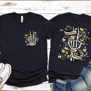 May include: Two black t-shirts featuring a skeleton hand graphic and floral design. The front shirt has a smaller graphic with the text "Good Energy is Contagious". The back shirt has a larger graphic with the text "Good Energy is Contagious".