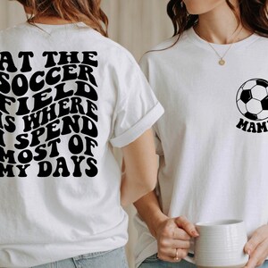 May include: Two white t-shirts with black text and a soccer ball graphic. The first shirt says "At the soccer field is where I spend most of my days". The second shirt says "Mama" and has a black and white soccer ball graphic.